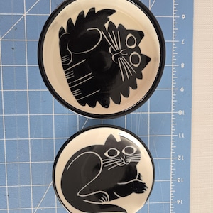 May include: Two ceramic dishes featuring black and white cat designs. The larger dish has a black cat with white eyes and a white background. The smaller dish has a white cat with black eyes and a black background.