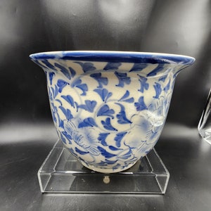 May include: A white ceramic bowl with a blue floral pattern. The bowl has a flared rim with a blue border. The floral design features blue flowers and leaves. The bowl is sitting on a clear acrylic stand.
