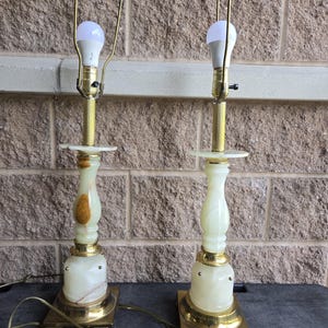 Puede incluir: Dos lámparas de mesa a juego con una base crema con aspecto de mármol y detalles en latón. Cada lámpara tiene una bombilla y un soporte de pantalla de metal dorado. Las lámparas están sobre una base cuadrada de latón.