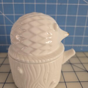 May include: White ceramic hedgehog trinket box with a tree trunk design. The lid is shaped like a hedgehog with a gold accent on the nose.