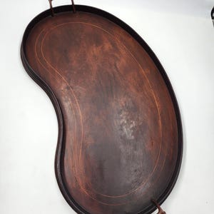 Könnte beinhalten: Ein großes, nierenförmiges Serviertablett aus Holz mit zwei dunkelbraunen Griffen. Das Tablett hat eine dunkelbraune Beize und einen hellbraunen Rand um den Rand.