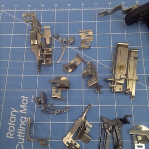 May include: A collection of silver-toned metal sewing machine parts, including a foot pedal, a needle plate, and a bobbin case. The parts are arranged on a blue cutting mat with a white grid pattern. The mat has the words "Rotary Cutting Mat" printed on it.