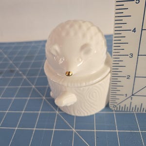 May include: White ceramic hedgehog figurine with a gold nose, sitting on a white ceramic log. The figurine is approximately 2 inches tall.
