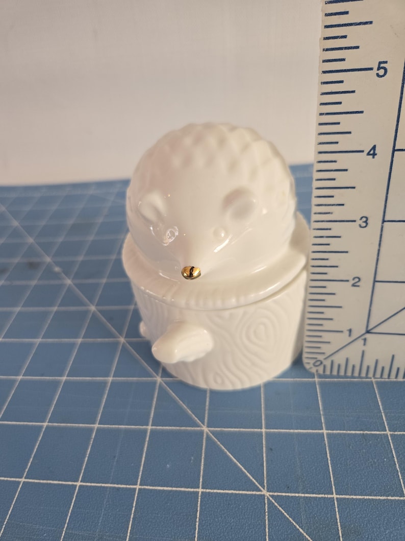May include: White ceramic hedgehog figurine with a gold nose, sitting on a white ceramic log. The figurine is approximately 2 inches tall.