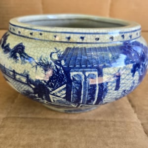 Può includere: Una ciotola rotonda in ceramica blu e bianca con un disegno a paesaggio. La ciotola presenta una glassa craquelé e una scena dettagliata di un edificio, alberi e un ponte. Il bordo ha una serie di piccoli segni blu scuro.