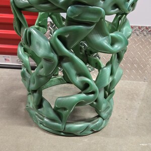 May include: A green, sculptural side table with a unique design. The table features a cylindrical shape with interwoven, flowing elements. The color is a solid, vibrant green, and the overall aesthetic is modern and artistic.