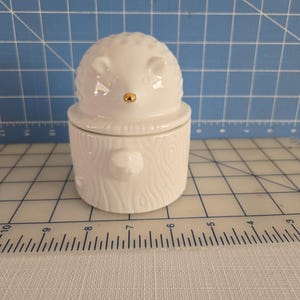 May include: White ceramic hedgehog shaped container with a gold nose. The container is shaped like a log with a wood grain texture.