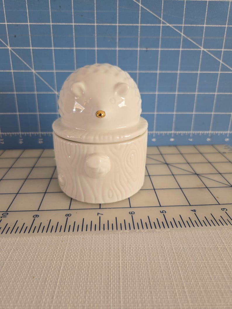 May include: White ceramic hedgehog shaped container with a gold nose. The container is shaped like a log with a wood grain texture.