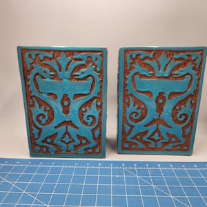 以下が含まれることがあります： スタイリッシュなドラゴンがデザインされた、ターコイズブルーのセラミック製ブックエンドのペア。ブックエンドは正方形で、立体的なデザインが施されています。