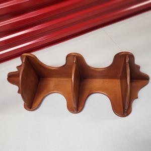 May include: A wooden shelf with three compartments, stained in a warm brown color. The shelf has a unique, curved design with decorative edges, suitable for displaying small items or decor.