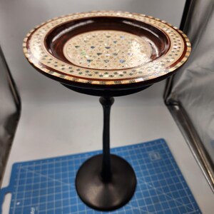 May include: A wooden pedestal with a round, inlaid top. The top is made of wood and mother of pearl and has a geometric pattern. The pedestal is black and has a round base.