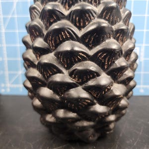 May include: A black ceramic pine cone ornament with a detailed textured surface.