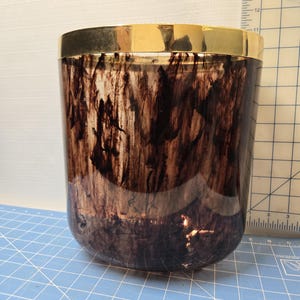 May include: A cylindrical container with a gold-colored rim. The body of the container has a dark brown, marbled pattern. The container is sitting on a blue surface with a grid pattern.