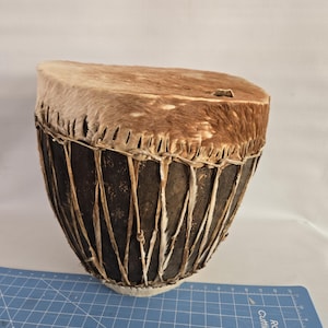 May include: A brown leather drum with a natural fur head. The drum is made of wood and has a rope-like design around the middle.