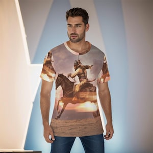 May include: A short-sleeved crew neck t-shirt featuring a full-front graphic print of a warrior in historical attire riding a dark horse across a desert landscape at sunset, with a warm orange and brown color palette throughout the entire garment.