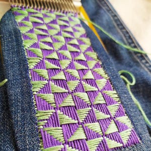 May include: Close-up of a purple and green woven tapestry with a geometric pattern. The tapestry is being woven on a loom.