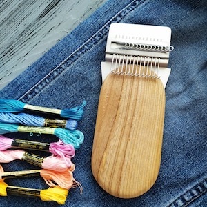 May include: A wooden loom tool with a metal frame and multiple hooks. The tool is on a blue denim background with colourful embroidery floss.