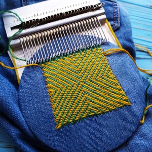Może przedstawiać: Biało-srebrne krosno z zieloną i żółtą nicią tworzące geometryczny wzór na niebieskim dżinsie. Dżins leży na niebieskiej drewnianej powierzchni. Krosno jest częściowo zmontowane.