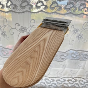 Può includere: Un telaio in legno con un pettine in metallo e una finitura in legno marrone chiaro. Il telaio è tenuto in una mano.