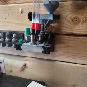 May include: A close-up of a reloading press mounted on a wooden wall. The press has a clear plastic tube with red and green accents. The press is labeled "Dillon Precision".
