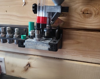 May include: A close-up of a reloading press mounted on a wooden wall. The press has a clear plastic tube with red and green accents. The press is labeled "Dillon Precision".