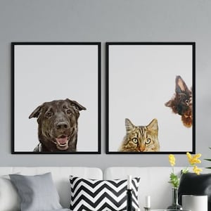 May include: Two framed prints of a dog and cat peeking out from behind the frames. The dog is brown and the cat is tabby. The prints are hanging on a white wall above a white sofa with grey and black cushions. A potted palm tree is on the right side of the image.