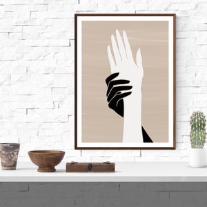Puede incluir: Una impresi&oacute;n de arte abstracto enmarcada que presenta dos manos, una negra y una blanca, superpuestas sobre un fondo beige.