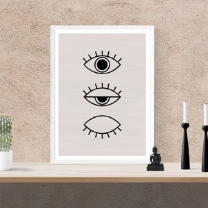 Puede incluir: Una impresi&oacute;n enmarcada con tres ojos negros estilizados sobre un fondo beige claro. La impresi&oacute;n se muestra en un estante con una peque&ntilde;a planta de cactus, una peque&ntilde;a estatua negra y dos candelabros negros.
