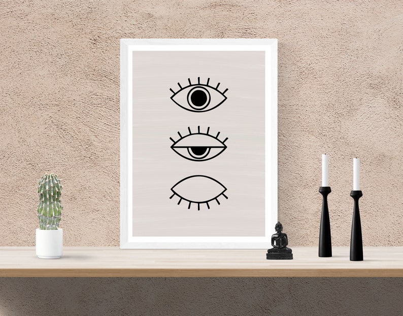 Puede incluir: Una impresi&oacute;n enmarcada con tres ojos negros estilizados sobre un fondo beige claro. La impresi&oacute;n se muestra en un estante con una peque&ntilde;a planta de cactus, una peque&ntilde;a estatua negra y dos candelabros negros.