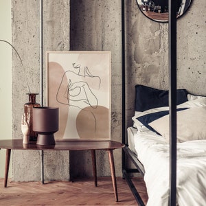 Puede incluir: Una impresi&oacute;n de arte de l&iacute;nea de una mujer desnuda con un fondo marr&oacute;n y beige. La impresi&oacute;n est&aacute; colgada en una pared de hormig&oacute;n sobre una mesa de caf&eacute; de madera.