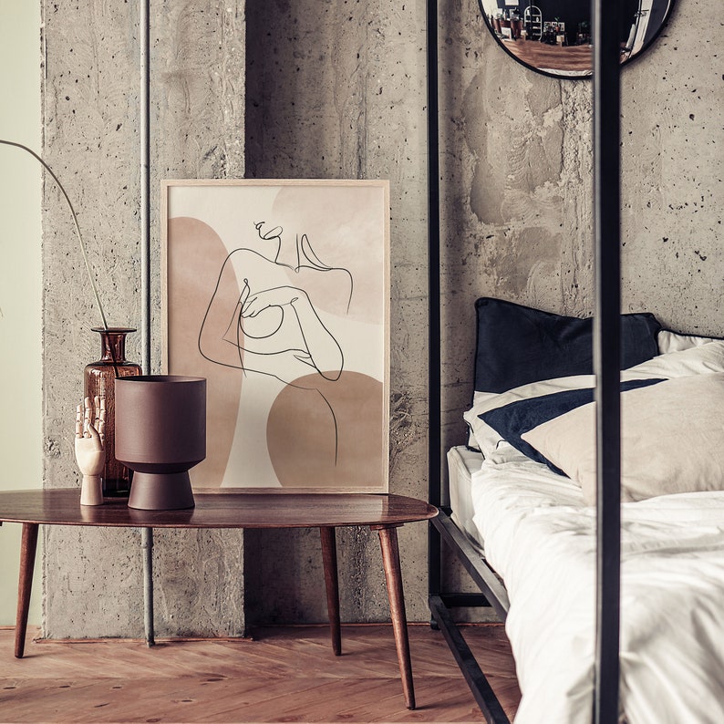 Puede incluir: Una impresi&oacute;n de arte de l&iacute;nea de una mujer desnuda con un fondo marr&oacute;n y beige. La impresi&oacute;n est&aacute; colgada en una pared de hormig&oacute;n sobre una mesa de caf&eacute; de madera.