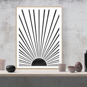 Op de afbeelding: Zwart-witte minimalistische kunstprint van een zonnestralenontwerp met een zwarte cirkel in het midden. De print is ingelijst in een licht houten lijst.