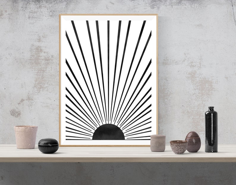 Op de afbeelding: Zwart-witte minimalistische kunstprint van een zonnestralenontwerp met een zwarte cirkel in het midden. De print is ingelijst in een licht houten lijst.