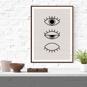 Puede incluir: Una impresi&oacute;n enmarcada con un dise&ntilde;o minimalista que presenta tres ojos estilizados en negro sobre un fondo beige claro.