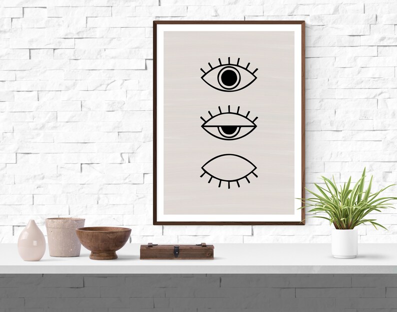 Puede incluir: Una impresi&oacute;n enmarcada con un dise&ntilde;o minimalista que presenta tres ojos estilizados en negro sobre un fondo beige claro.