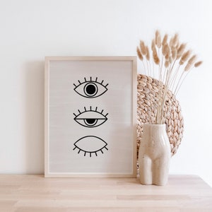 Puede incluir: Una impresi&oacute;n enmarcada con tres ojos estilizados negros sobre un fondo blanco. La impresi&oacute;n est&aacute; colgada en una pared blanca sobre una mesa de madera. Un jarr&oacute;n con hierbas secas est&aacute; sobre la mesa frente a la impresi&oacute;n.