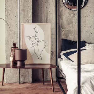 Może przedstawiać: Minimalistyczny wydruk sztuki liniowej przedstawiający dwie postaci w miękkiej r&oacute;żowo-beżowej kolorystyce. Wydruk jest oprawiony i wisi na ścianie nad drewnianym stolikiem kawowym.