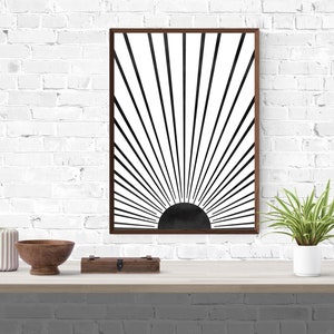 Op de afbeelding: Een minimalistische zwart-witte zonsondergangprint in een bruin frame. De print is op een witte achtergrond met zwarte lijnen die uit een zwarte cirkel in het midden stralen.