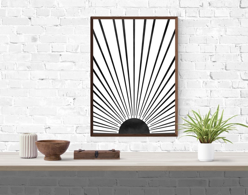Op de afbeelding: Een minimalistische zwart-witte zonsondergangprint in een bruin frame. De print is op een witte achtergrond met zwarte lijnen die uit een zwarte cirkel in het midden stralen.