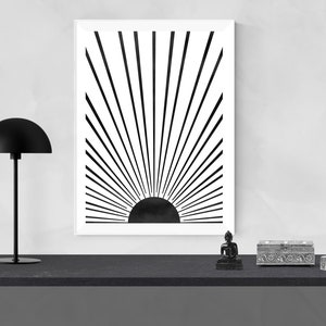 Op de afbeelding: Zwart-witte minimalistische kunstprint van een zon met stralen die er vanaf komen. De print is ingelijst en hangt aan een witte muur boven een zwarte plank met een zwarte lamp en decoratieve voorwerpen.