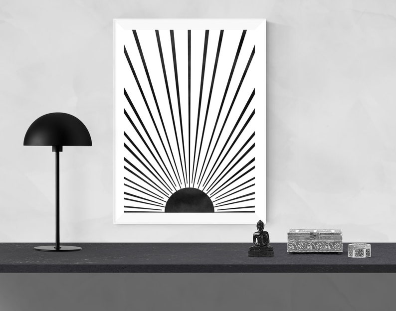 Op de afbeelding: Zwart-witte minimalistische kunstprint van een zon met stralen die er vanaf komen. De print is ingelijst en hangt aan een witte muur boven een zwarte plank met een zwarte lamp en decoratieve voorwerpen.
