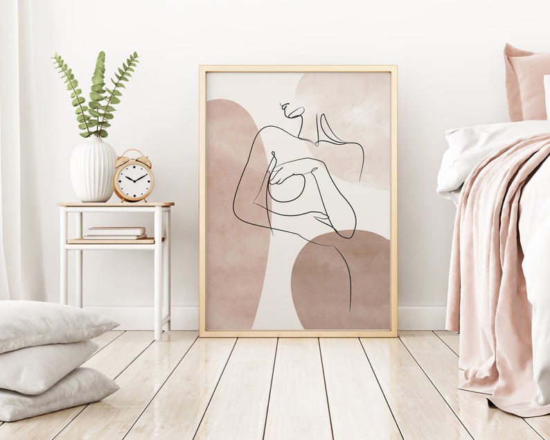 Puede incluir: Un dibujo de l&iacute;nea minimalista de una persona con un cuerpo desnudo, sobre un fondo de formas abstractas en tonos rosa y marr&oacute;n. La obra de arte est&aacute; enmarcada en un marco dorado.