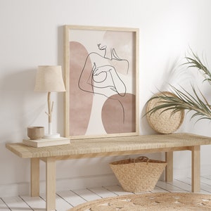 Puede incluir: Un banco de madera con un asiento tejido y una cesta de mimbre debajo. Una impresi&oacute;n enmarcada de un dibujo lineal de una mujer con formas abstractas en tonos rosa y marr&oacute;n apagados cuelga de la pared detr&aacute;s del banco.