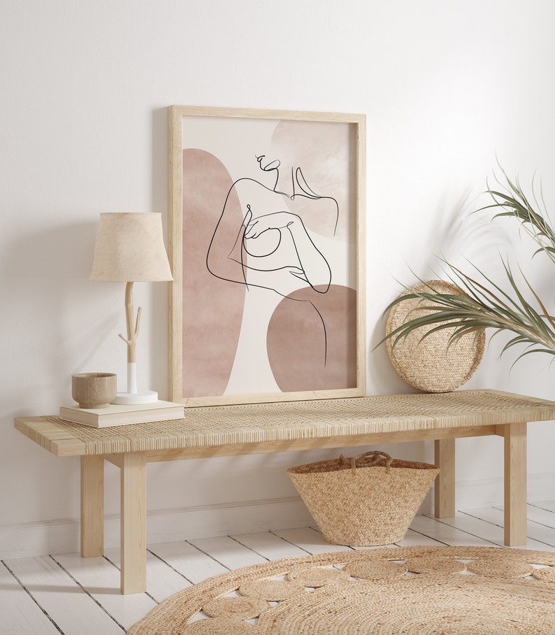 Puede incluir: Un banco de madera con un asiento tejido y una cesta de mimbre debajo. Una impresi&oacute;n enmarcada de un dibujo lineal de una mujer con formas abstractas en tonos rosa y marr&oacute;n apagados cuelga de la pared detr&aacute;s del banco.