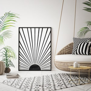 Op de afbeelding: Zwart-witte geometrische zonsondergangprint in een zwart frame dat aan een witte muur hangt boven een wit-zwart gepatroneerd tapijt. Een hangende rieten stoel met zwart-wit gestreepte kussens staat op de voorgrond.