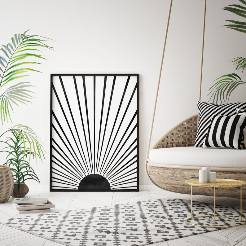 Op de afbeelding: Zwart-witte geometrische zonsondergangprint in een zwart frame dat aan een witte muur hangt boven een wit-zwart gepatroneerd tapijt. Een hangende rieten stoel met zwart-wit gestreepte kussens staat op de voorgrond.