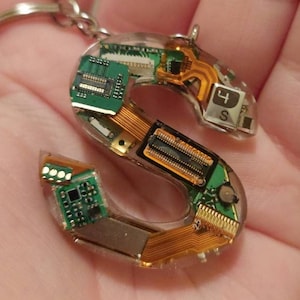 May include: A clear resin keychain in the shape of the letter "S", filled with electronic components. The components include green circuit boards, gold connectors, and small black chips. A silver keyring is attached at the top.