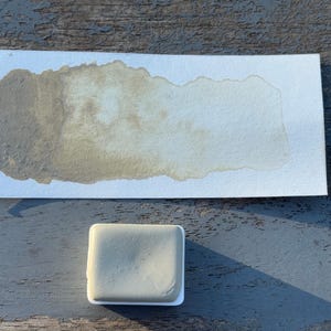 May include: A watercolor paint swatch on white paper with a rectangular paint block. The paint swatch is a blend of light and dark olive green hues. The paint block is a light olive green color.