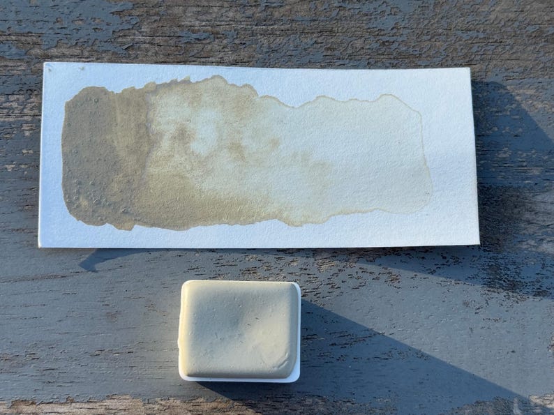 May include: A watercolor paint swatch on white paper with a rectangular paint block. The paint swatch is a blend of light and dark olive green hues. The paint block is a light olive green color.