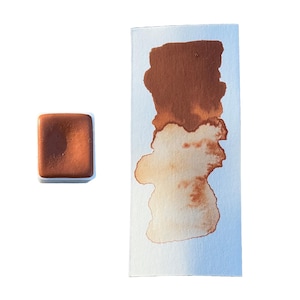 May include: A watercolor paint pan with a brown pigment. The paint is a deep brown color with a hint of orange. The pan is made of white plastic and has a square shape.
